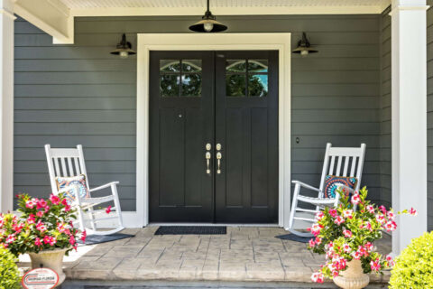 A front porch of a home with double glass exterior doors, two white rocking chairs and flowers in planters